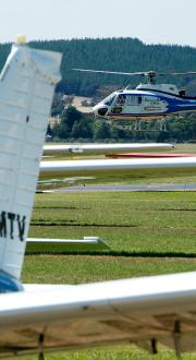 The wing and tail of a small aircraft at an airfield is in the foreground. In the background there is another small airplane and a hovering helicopter. 