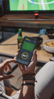 Man sitting on a couch watching a soccer match, holding a phone in his hand with a betting site open 