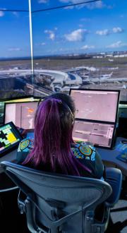 Photograph of an air traffic controller in a control tower managing multiple digital screens displaying radar, flight paths, and airport layout. Large windows provide a panoramic view of an airport terminal with several parked airplanes.