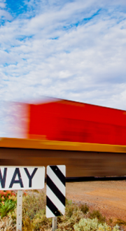 A stop and look for trains sign in the foreground with a blurry green, red and blue rail train in the background driving over a level crossing on a dirt road. 