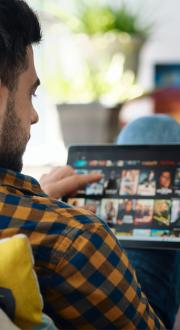 Man sitting on couch scrolls through titles available on a streaming service on an Ipad.
