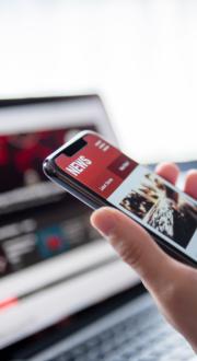 A person holding a mobile phone with a news website open, a laptop sits in the background with a website open.