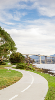 Shared bike and walking path, running alongside a road, hugging a body of water, with a bridge and mountains in the distance.