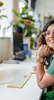 Woman seated at a desk in a modern office workspace, wearing glasses and a green outfit. A computer monitor, keyboard and indoor plants are visible, with a colleague and office area softly blurred in the background.
