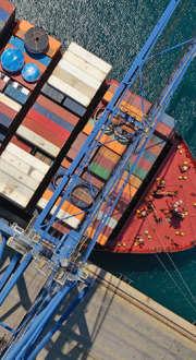 Looking down on a cargo ship with shipping containers, docked at the port next to gantry cranes. 