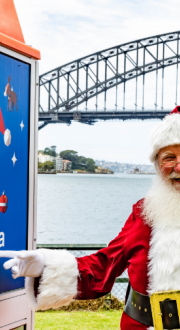 Santa pointing to a Telstra payphone with ‘Call Santa here’ printed on it in front of Sydney harbour.