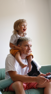 Father sitting on a couch playing video games with child sitting on his shoulders