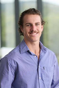 Close up portrait photo of a smiling Declan Tancred wearing a blue long sleeve collared shirt. Declan has brown hair and a moustache.
