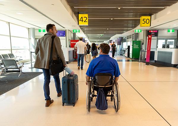 Image of two people moving through an airport. The person on the right is using a wheelchair and the person on the left is walking with a suitcase. 